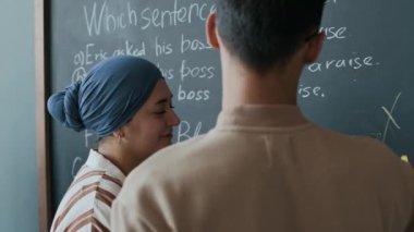 Erkek İngilizce öğretmeni, karatahta üzerindeki cümle düzeltme egzersizlerini kontrol ediyor ve ders sırasında bayan Müslüman öğrencilere hataları anlatıyor.