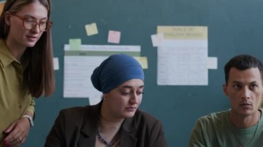 Orta boy beyaz kadın öğretmen, kültürler arası bir grup öğrencinin masada oturup İngilizce dil bilgisi öğrenmesine yardım ediyor.