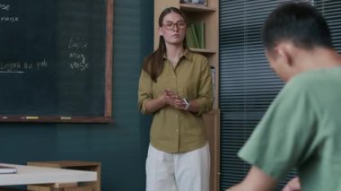 Orta boy beyaz kadın İngilizce öğretmeni karatahtaya dil bilgisi kuralları yazıyor ve modern sınıflarda ders verirken çift ırklı erkek öğrenci notlarını kontrol ediyor.