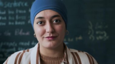Genç, yetişkin Müslüman bir öğrencinin, tahtaya tebeşirle yazılmış İngilizce egzersizle fotoğraf çektirdiği orta boy bir portresi.