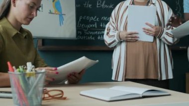 Beyaz tenli kadın İngilizce öğretmeninin beliyle yazı tahtasının yanında oturması ve ders sırasında öğrenci raporlarını kontrol etmesi.