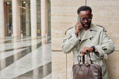 Modern iş binasının önünde duran, elinde deri bir çantayla telefonla konuşan, trençkot giyen bir adam.