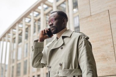 Modern bir binanın önünde şık bir trençkot ve güneş gözlüğü takmış bir adam dışarıda telefonla konuşuyor.