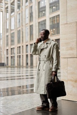 Trençkotlu bir adam modern kentsel alanda duruyor. Elinde evrak çantasıyla cep telefonuyla konuşuyor. Arkaplanda geniş pencere ve sütunlara sahip çağdaş mimari