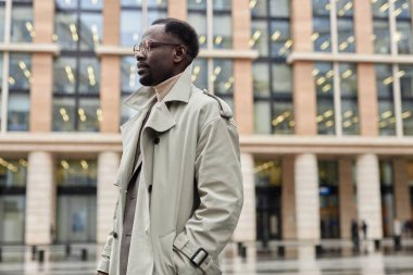 Trençkot giyen bir adam modern cam binanın yanında yürüyor. Turuncu aksanlı, arka planda çok sayıda ışıklı pencere var.