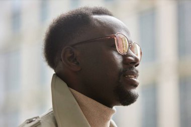 Şık gözlüklü ve bej ceketli bir adam, modern binalarla şehir merkezinde dikiliyor, düşünceli bakışlarını başka yöne çeviriyor.