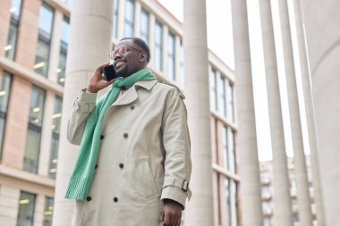 Açık renk ceket giyen ve yeşil atkı takan bir adam modern ofis binasının dışında telefonla konuşurken arka planda uzun sütunlar ve cam pencerelerle gülüyor.