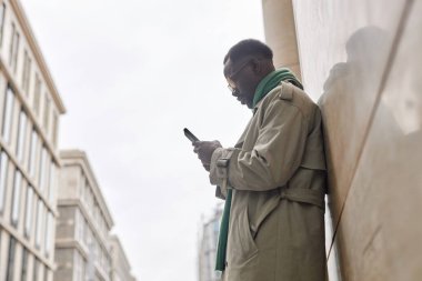 Akıllı telefonuna bakarken binanın yanında duran Afro-Amerikalı bir adam. Arka planda yüksek binalar olan bir ceket ve eşarp giyiyor.