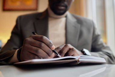 Person writing in notebook during business meeting, focusing on taking detailed notes while sitting near window with natural light illuminating surroundings clipart