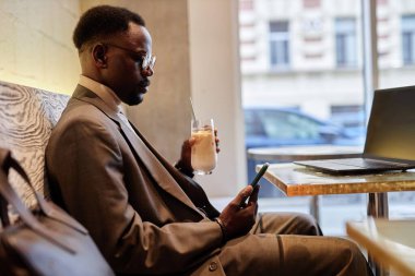 Kafede oturan takım elbiseli Afro-Amerikalı bir adam elinde cep telefonu ve içecek tutarak ekrana bakıyor. Çalışma alanı dizüstü bilgisayar ve bir bardak milkshake görünür