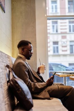 Afro-Amerikalı bir adam kafede oturuyor, aşağı bakarken içki içiyor. Elindeki içecekle rahatça oturarak, rahat bir bankta rahatlamanın tadını çıkarıyorsun.