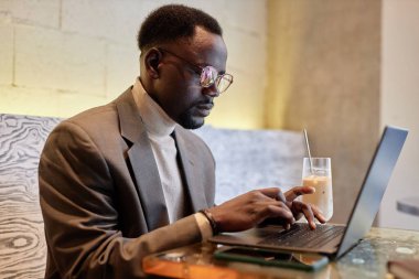 Afro-Amerikan erkek, rahat bir kafede dizüstü bilgisayarda çalışıyor, şık gözlükler takıyor ve temiz bir bardaktan içkisini yudumluyor, daklara konsantre oluyor.