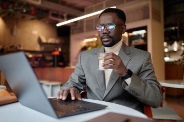 Afro-Amerikalı bir adam gözlük takıyor ve laptopla oturmuş kafede kahve içiyor, işine dalmış, modern çevrede hareketli bir ortam ve konsantrasyon gösteriyor.