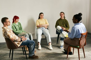 Çeşitli genç yetişkinler çemberde otururken profesyonel terapist rahat bir odada sohbet ve aktivitelere katılan katılımcıları yönetiyor.