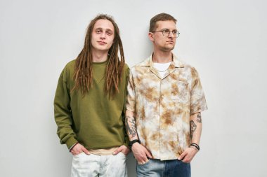 Biri yeşil kazaklı, diğeri gözlüklü ve kollarında dövmeleri olan baskılı gömlekli iki adam.