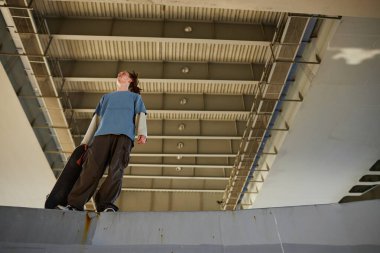 Endüstriyel bir köprünün altında kaykayı tutan ve yukarı bakan biri. Işıklandırma köprünün yapısal detaylarını vurguluyor