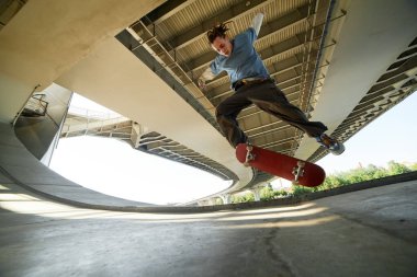 Genç erkek kaykaycı şehir köprüsü altında havanın ortasında dinamik hareketi ve kentsel ayarı yakalayan beton yapılarla bir numara yapıyor.