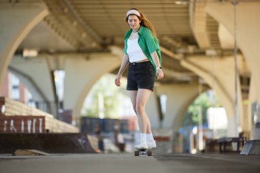Şehir köprüsünün altında kaykay yapan kişi günlük kıyafet ve saç bandı takıyor. Mimari yapılar ve dinamik atmosferi yansıtan canlı çevreyle dolu sahne