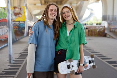 Kaykaylı genç çift kameraya gülümserken şehir köprüsünün altında poz veriyor. Grafiti ve beton yapıların yer aldığı arka plan canlı kentsel atmosfer yaratıyor