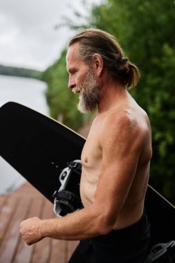 Sakallı, aktif, elinde sörf tahtası olan yaşlı bir adamın portresi. Gölün kıyısındaki tahta iskelede duran yaz sporlarının keyfini çıkarmaya hazır.