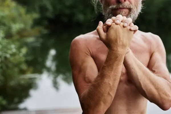 stock image Cropped shot of sportive shirtless senior man stretching and doing warm up for hands and arm muscles standing by lake in forest copy space