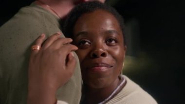 Genç, gülümseyen zenci bir kadının gece açık havada dans ederken başını erkeğin omzuna koyduğu orta boy yakın çekim.