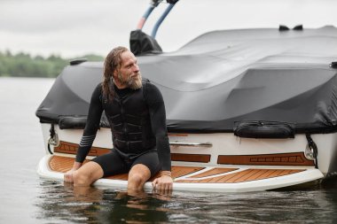 Göl ve açık havada yüzmenin keyfini çıkardıktan sonra dalış kıyafeti giyen ve teknenin arkasında oturan son sınıf sporcusunun samimi portresi.