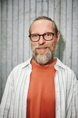 Gözlüklü, sakallı, gülümseyen bir adamın dikey portresi ve metal duvarlara dayalı bir kamera görüntüsü.