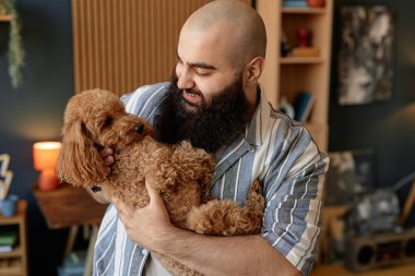 Gülümseyen adam rahat oturma odasında yumuşacık kahverengi köpeğe sarılıyor sıcak dekor ve kişisel eşyalarla çevrili memnuniyet ve şefkat portresi.