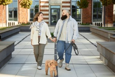 Çiftler, köpekleri tasmalı olarak dışarıda el ele yürüyorlar. Güneşli bir günde, arka planda modern binalar bulunan günlük giysiler içinde, mutlu mesut gülümseyip birbirlerine bakıyorlar.