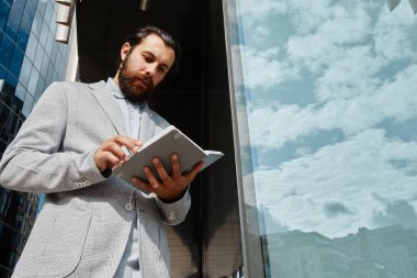 Sakallı adam, açık havada cam binanın yanında dururken tablet üzerindeki verileri analiz ediyor. Mavi gökyüzünü ve camdaki bulutları yansıtıyor.