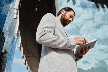 Modern cam binanın önünde duran gri takım elbiseli sakallı adam, iş için tablet kullanıyor. Şehir manzarası ve pencerelere yansıyan bulutlar