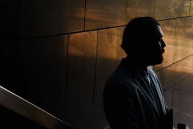 Sönük bir koridora bakan bir kişinin silueti profilinde gölgeleri vurguluyor, gizemli ve yansıtıcı bir ortam yaratıyor.