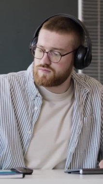 Ofiste bilgisayar başında çalışırken kulaklık takan genç beyaz programcı akıllı telefondan bildirimi kontrol ediyor.