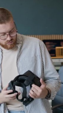 VR kulaklığı tutan genç beyaz bilişim uzmanının orta boy portresi kameraya bakıyor.