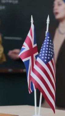 Beyaz kadın İngilizce öğretmeni ve genç yetişkin Müslüman kadın dikey olarak tahtada duruyor ve ders sırasında Amerikan ve İngiliz bayrakları seçici bir odak noktasında masada tartışıyorlar.