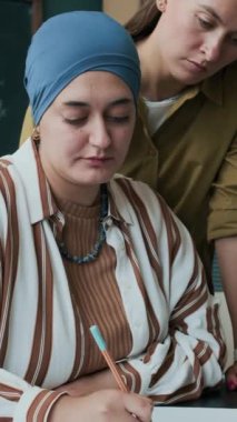 Kafkas kadın öğretmenin sınıfta genç Müslüman bir kadının kitap yazmasına yardım ettiği dikey bir fotoğraf.