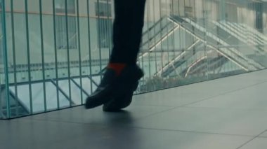 Siyah üniformalı bir erkek ofis çalışanının tuhaf dans figürleri sergilediği, çağdaş iş merkezinin cam korkuluklarına dayandığı bir sahne.