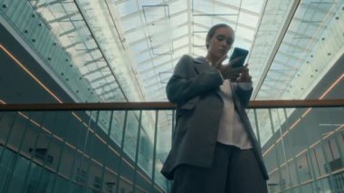 Düşük açılı, gri takım elbiseli beyaz bir iş kadını modern iş merkezinin cam parmaklıklarına yaslanırken, akıllı telefonuyla, sonra da eksantrik hareketlerle çağdaş dans etmeye başlarken.