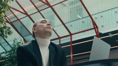 Beyaz balıkçı yaka ve siyah takım elbiseli, beyaz balıkçı yakalı beyaz bir adam gözbağını çıkarıp masa başında otururken ve modern kapalı alanda dizüstü bilgisayar üzerinde çalışırken çağdaş dans hareketleri yapmaya başlıyor.