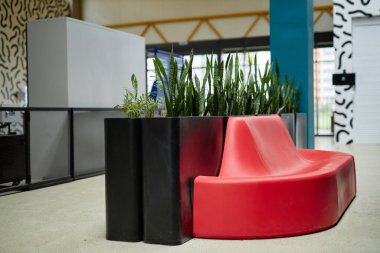 Modern ofis merkezi koridorunda şirket çalışanları için minimalist tasarım ve yeşillik içeren canlı kırmızı renkli rahat koltuklar var.