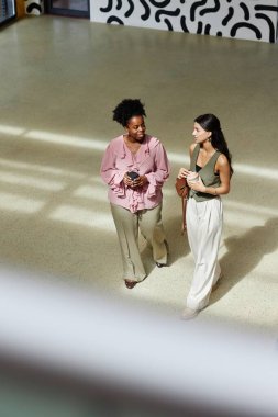 Vertical shot of fashionable Black businesswoman at walking meeting with female colleague discussing things over cup of coffee in office center, copy space clipart