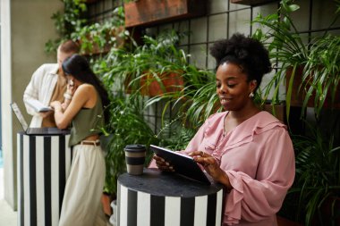 Orta büyüklükte gülümseyen bir iş kadını, yeşil duvarlı modern ofis merkezinin kafesinde çalışırken ayakta duran dijital tableti kullanırken,