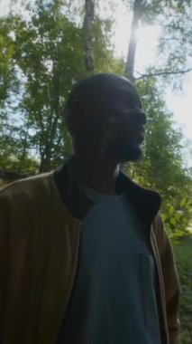 Kameraya poz veren genç siyahi erkek turistin dikey portresi yemyeşil ormanda gün ışığı altında duruyor.