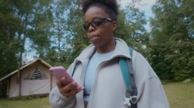 Güneş gözlüklü ve tüylü ceketli genç siyahi kadın akıllı telefon kullanırken gülümsüyor, çadır benzeri kulübenin yanında duruyor doğal orman ortamında çadır çatısı olan, tek başına yürüyüş deneyimi yaşıyor.