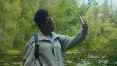 Afrika kökenli Amerikalı kadın yürüyüşçünün güneş gözlüğü ve sırt çantası takıp sakin göl kenarındaki yemyeşil ormanda selfie çekmek için durduğu orta boy bir fotoğraf.