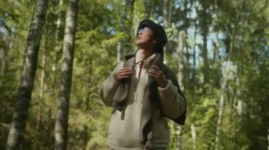 Sırt çantası ve kova şapkalı genç Asyalı erkek yürüyüşçünün, yemyeşil ormandaki uzun yeşil ağaçların arasında dikilirken orta açı görüntüsü.