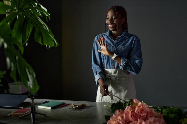 Minimal portrait of young African American woman as florist creating bouquet and filming process with smartphone for social media waving at camera copy space