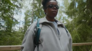 Genç Afro-Amerikan kadın yürüyüşçünün orta boy portresi, güneş gözlüklü, gür ormandaki ahşap köprüde duran ve yaz zamanı yürüyüş macerasında kameraya bakan.