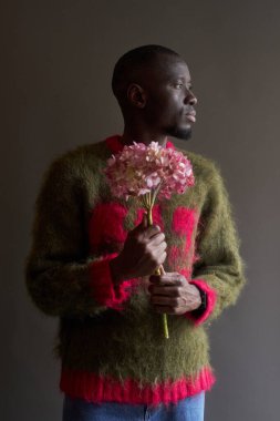 Çiçekli pembe çiçekleri tutan ve yumuşak tüylü süveter giyen yumuşak erkekliğin dikey moda portresi.